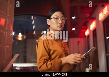 Girovita ritratto di moderna donna d'affari asiatica tenendo tablet e guardando la fotocamera mentre si posa all'aperto, copiare spazio Foto Stock