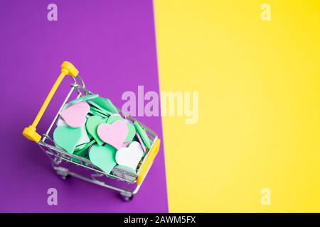 Primo piano di un carrello in miniatura pieno di cuori colorati su uno sfondo giallo e viola a due tonalità. San Valentino, primavera, amore, sconti e. Foto Stock