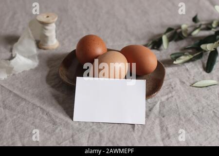 Primavera mockup scena. Messaggio di saluto vuoto, biglietto da visita sul tavolo. Pasqua, design fattoria. Foto in borsa dallo stile naturale femminile. Uova di pollo e ramo di olive Foto Stock