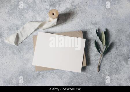 Cancelleria femminile per matrimoni, scena di simulazione desktop. Biglietto d'auguri bianco, busta artigianale con ramificazione di olive e nastro di seta.sfondo tavolo Grunge Foto Stock
