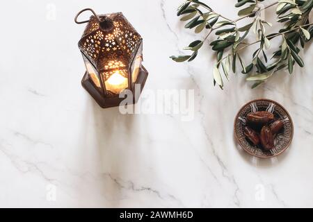 Biglietto d'auguri Ramadan Kareem, invito. Piatto in bronzo con datteri di frutta, rami d'oliva e luminosa lanterna marocchina su tavolo in marmo bianco. Iftar Foto Stock