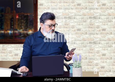 Un uomo d'affari maturo e di successo con occhiali da vista che lavora su un computer portatile mentre si utilizza il telefono in ufficio moderno Foto Stock