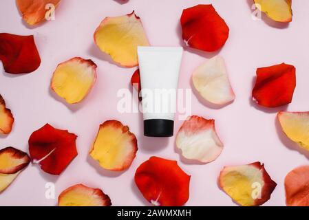Tuba con crema idratante su sfondo pastello rosa tra petali di rosa rosso e arancione. Il concetto di cura della pelle, cura anti-invecchiamento, ingredienti naturali in cos Foto Stock