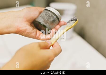 Spazzolino da denti in bambù e dentifricio al carbone in vaso di vetro nelle mani, pulizia dei denti del mattino. Concetto di bagno zero rifiuti. Riciclare e riutilizzare. Avampiede in legno Foto Stock