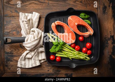 Bistecche di pesce crudo e verdure su una padella grigliata in ghisa su un rustico tavolo in legno, vista dall'alto. Mangiare sano, cucinare sano concetto di cibo Foto Stock