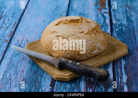 Pane fresco rotondo lievitato fatto in casa sul tagliere. Un vecchio coltello con un manico di legno è sdraiato davanti ad esso. Bellissimo sfondo rustico. Foto Stock