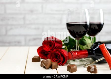 Concetto di San Valentino. Due bicchieri di vino, bottiglia di vino, cavatappi, rose rosse e cuori di cioccolato su un tavolo di legno bianco con spazio copia per il testo. Foto Stock