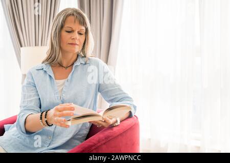 Donna matura che legge un libro nel suo appartamento urbano Foto Stock