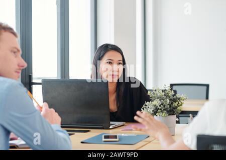 Grazioso giovane sviluppatore di software asiatico seduto al tavolo con il laptop aperto e l'ascolto di idee dei product manager Foto Stock