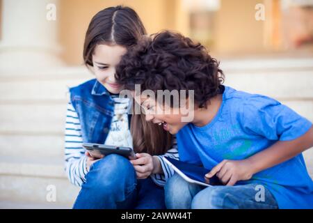 Due bambini ragazzo e ragazza che giocano a videogame su tablet all'aperto. Concetto di dipendenza da gadget e bambini Foto Stock