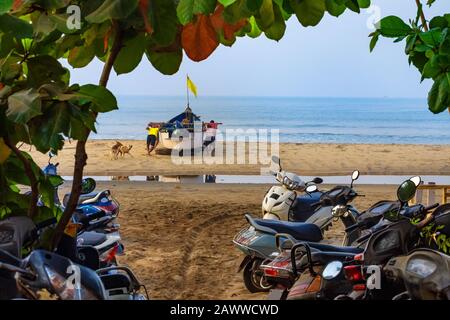 Un gruppo di pescatori spinge una barca tradizionale verso il mare. Tipico paesaggio mattutino di un paese esotico. Parcheggio in primo piano per scooter. Arambol, G. Foto Stock