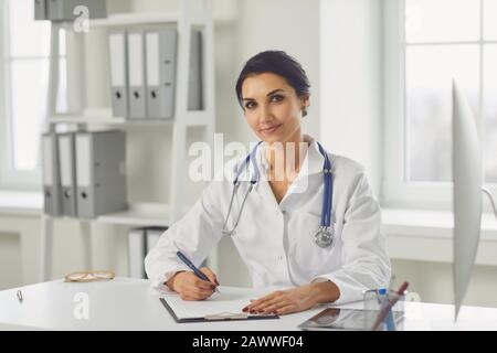 Il medico sicuro della donna pediatra scrive in un portadocumenti seduto ad un tavolo in un ufficio bianco dell'ospedale Foto Stock