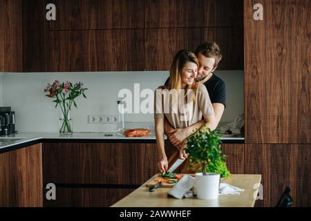 L'uomo cerca l'attenzione, abbracciando la sua ragazza mentre sta affettando le verdure Foto Stock