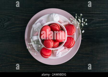 Sfondo di San Valentino con macaroni rosa a forma di cuore e marshmallows su un piatto. Sfondo di legno scuro. Disposizione piatta. Foto Stock