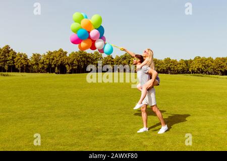 Giovane e simpatico uomo che cammina nel parco e che porta indietro la sua ragazza con palloncini Foto Stock