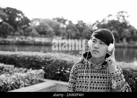 Giovane ragazzo asiatico teenage indossa le cuffie mentre si rilassa al parco Foto Stock