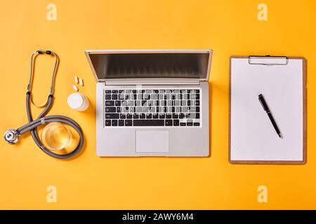 Laptop, carte, stetoscopio, farmaci e vetro d'acqua su sfondo giallo, piatto orizzontale scatto Foto Stock