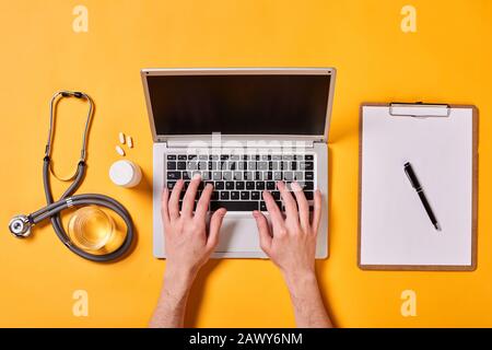 Laptop, mani di donna che lavora su di esso, carte, stetoscopio, farmaci e vetro di acqua su sfondo giallo, orizzontale piatto scatto lay Foto Stock