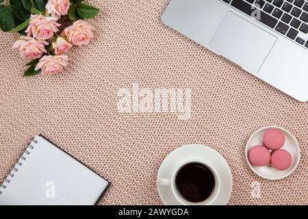 Lavoro da casa concetto. Moderno spazio di lavoro femminile, vista dall'alto. Laptop, caffè, torte, rose, taccuino su coperta a maglia, spazio di copia, piatto lay. Deskto Foto Stock