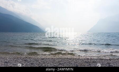 Vista panoramica sul lago di Garda da una spiaggia di ciottoli al sole e luce nebulosa. Rilassante paesaggio acquatico di onde luminose sulla superficie di un Foto Stock