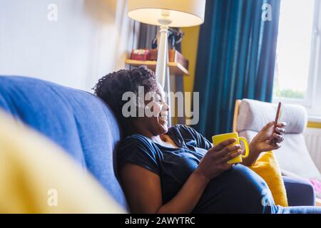 Donna incinta felice rilassarsi sul divano con tè e smartphone Foto Stock