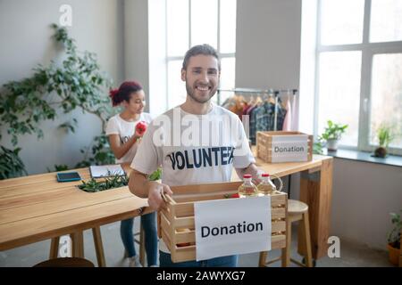 Giovane uomo felice che tiene la scatola di donazione, ragazza al tavolo. Foto Stock