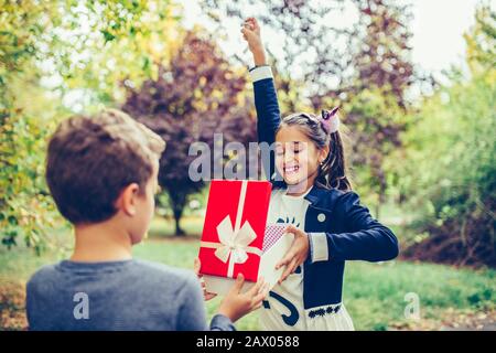 Felice bambino bambino dà alla ragazza sorridente una scatola regalo, facendo una sorpresa. Felice bambina gioisce dono che ha ricevuto dal suo migliore amico Foto Stock