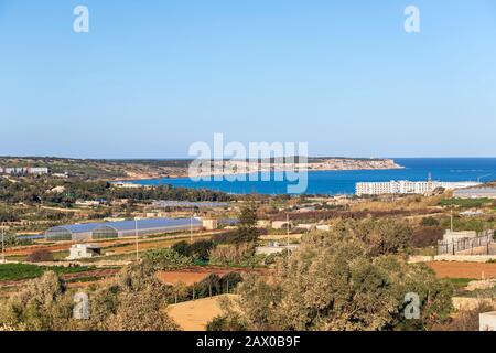 Campagna rurale dell'isola maltese con vegetazione secca, campi coltivati, terra arata e acque blu della baia mediterranea sullo sfondo, Malta Foto Stock