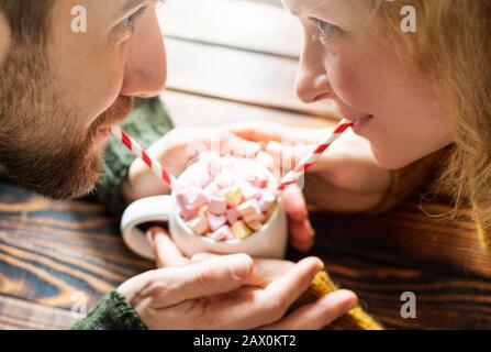 Giovane coppia bevendo cioccolata calda con marshmallows a forma di cuore usando cannucce da bere di carta. Data di San Valentino Foto Stock