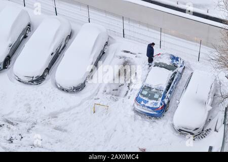 Bucarest, Romania - 6 febbraio 2020: L'uomo libera la neve dalla sua auto con una spazzola, su una fila con auto parcheggiata coperta da un nuovo strato di neve. Alto ang Foto Stock