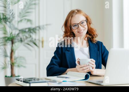 Indoor shot di femminile editor funziona su articolo, utilizza computer portatile e internet wireless, controlla le ultime notizie, tiene tazza di caffè o tè, circondato Foto Stock