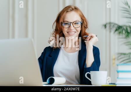 Felice redhead curly donna lavora freelance, utilizza computer portatile, scrive le note in blocco note, bevande caffè, indossa occhiali ottici, essere di buon umore Foto Stock