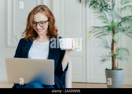 Ottimista capelli rossi giovane riuscito freelancer femminile lavora a distanza nello spazio di coworking, fa ricerca, fa corsi online, siede in poltrona, chiave Foto Stock