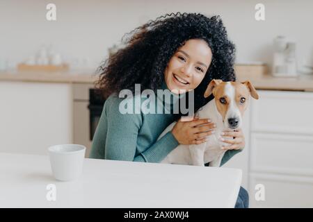 Adorabile donna sorridente abbraccia il suo cane preferito, esprime la cura e l'amore, buon attitiude ad animale domestico, pone contro l'interno della cucina, beve beve beve beve aromatiche Foto Stock