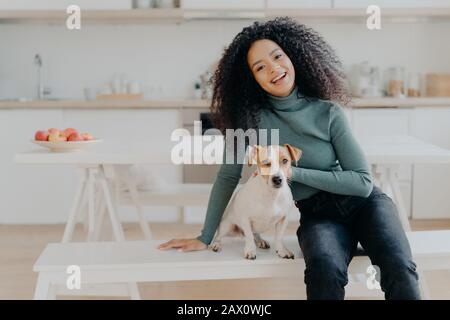 Il proprietario dell'animale domestico femminile esprime l'amore al cane, vestito in tleneck casual e jeans, siede alla panchina bianca contro l'interno accogliente della cucina, spende il tempo libero a. Foto Stock