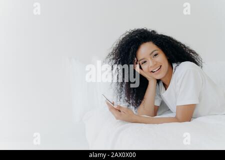 Overjoyed rilassato curly giovane afroamericana donna gode di tempo libero a casa, si trova a letto con moderno cellulare, utilizza il telefono in camera da letto, invia il testo m Foto Stock