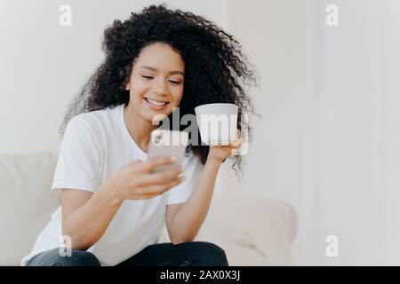 Tranquilla curly donna scorrimento social network tramite moderno cellulare, orologi video divertenti, guarda foto in social network, bevande caffè, pose on Foto Stock