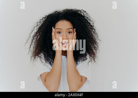 Foto di sorpresa donna allegra con capelli Afro, mantiene entrambe le mani sulle guance, ha bellezza naturale, apre la bocca, non può credere in notizie eccitanti, wea Foto Stock