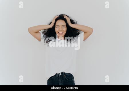 La foto della donna dai capelli ricci e allegra ride felicemente, è divertente, vestita di maglietta bianca e jeans, sorride ampiamente, isolata su sfondo bianco. Peo Foto Stock