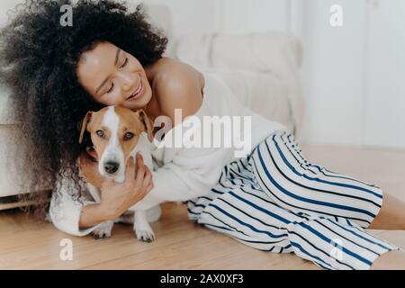 Affettuosa giovane donna abbraccia cane con amore e cura, mantiene gli occhi chiusi dal piacere, sorride dolcemente, ha sana pelle scura, posa sul pavimento, accarezzando un Foto Stock