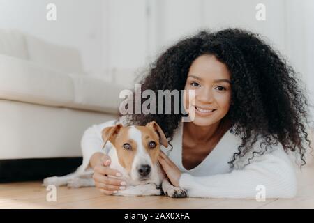 Bella ragazza con capelli afro, si trova a terra con cane, esprime piacevoli emozioni, dispone in soggiorno vicino al divano, ha acquistato animale domestico in nuovo appartamento. Donna h Foto Stock