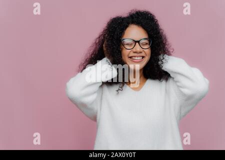 Ritratto di gioiosa donna afroamericana dall'aspetto piacevole copre entrambe le orecchie con le mani, ascolta musica forte, sorride ampiamente, indossa occhiali e bianco w Foto Stock