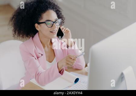 Felice giovane afroamericana donna discute notizie di affari, punti con penna a schermo sul computer, colloqui via telefono cellulare moderno, occupato con lavoro di carta, w Foto Stock