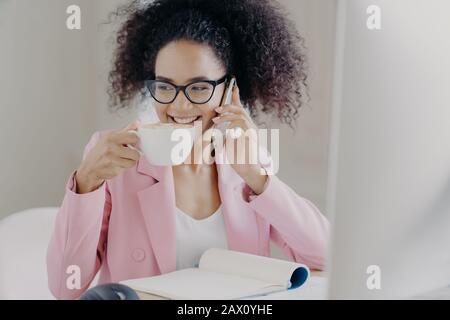 L'immagine ritagliata della giovane ragazza africana africana felice americana fa la telefonata, bevande latte aromatico o espresso, pose in ufficio interno, ha sorriso piacevole Foto Stock