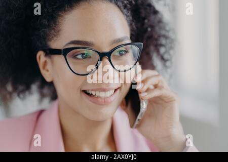 Headshot di bella donna sorridente pelle scura indossa occhiali ottici, tiene cellulare moderno, ha curato la carnagione, parla di futuro investm Foto Stock