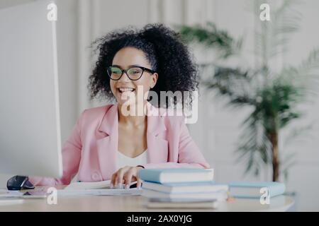 Scatto orizzontale del freelancer africano allegro americano browses del Internet sul calcolatore, messo a fuoco allo schermo, indossa gli occhiali ottici ed il costume ufficiale rosato Foto Stock