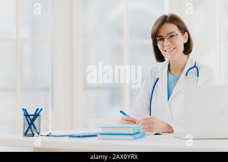 Medico specialista indossa abito bianco con stetoscopio, lavora in ospedale, prende appunti, posa in proprio cabinet, si siede al tavolo con computer portatile in offi Foto Stock