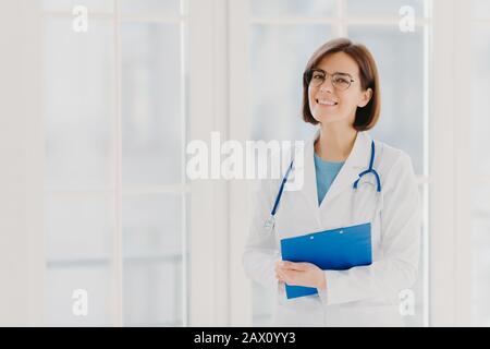 Donna esperta pediatra stand con appunti in armadietto, indossa cappotto medico bianco con stetoscopio, dà eccellente trattamento medico, ha hap Foto Stock