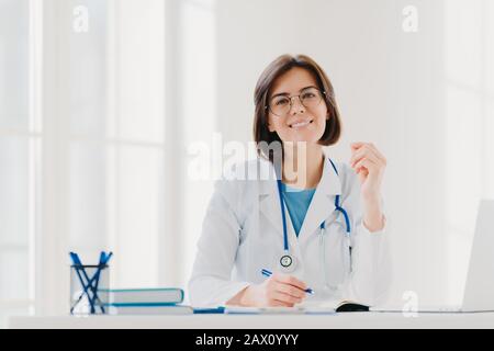 Giovane donna medico scrive su carta in clinica, creats prescrizione per il paziente, fa la documentazione in ospedale, indossa medico bianco abito, si siede in modo accogliente Foto Stock