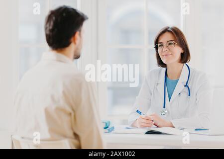 Il medico e il paziente discutono qualcosa, si siedono al tavolo in clinica. Il cardiologo femminile negli occhiali fornisce la diagnostica di consultazione medica, consiglio per l'uomo come Foto Stock